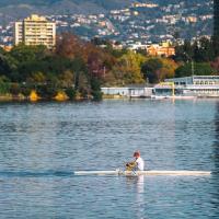 A view of Lake Merrit, with a man rowing across the water in the foreground, and trees and buildings reflecting on the water from the shore.