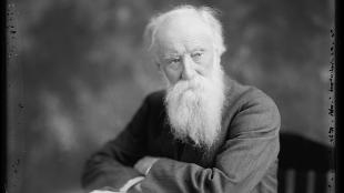 Black and white photo of American author John Burroughs, seated at a table with an open book before him