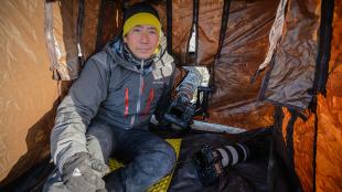 Photographer and author Noppadol Paothong, dressed for cold weather, sits inside a tent with openings for his cameras used for photographing wildlife.