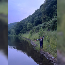 Woman wearing black stands on the edge of a lake, with arms stretched out